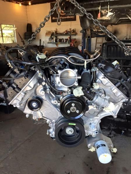 A car engine suspended by a chain in a garage, featuring a black pulley system and a white oil filter at the bottom.
