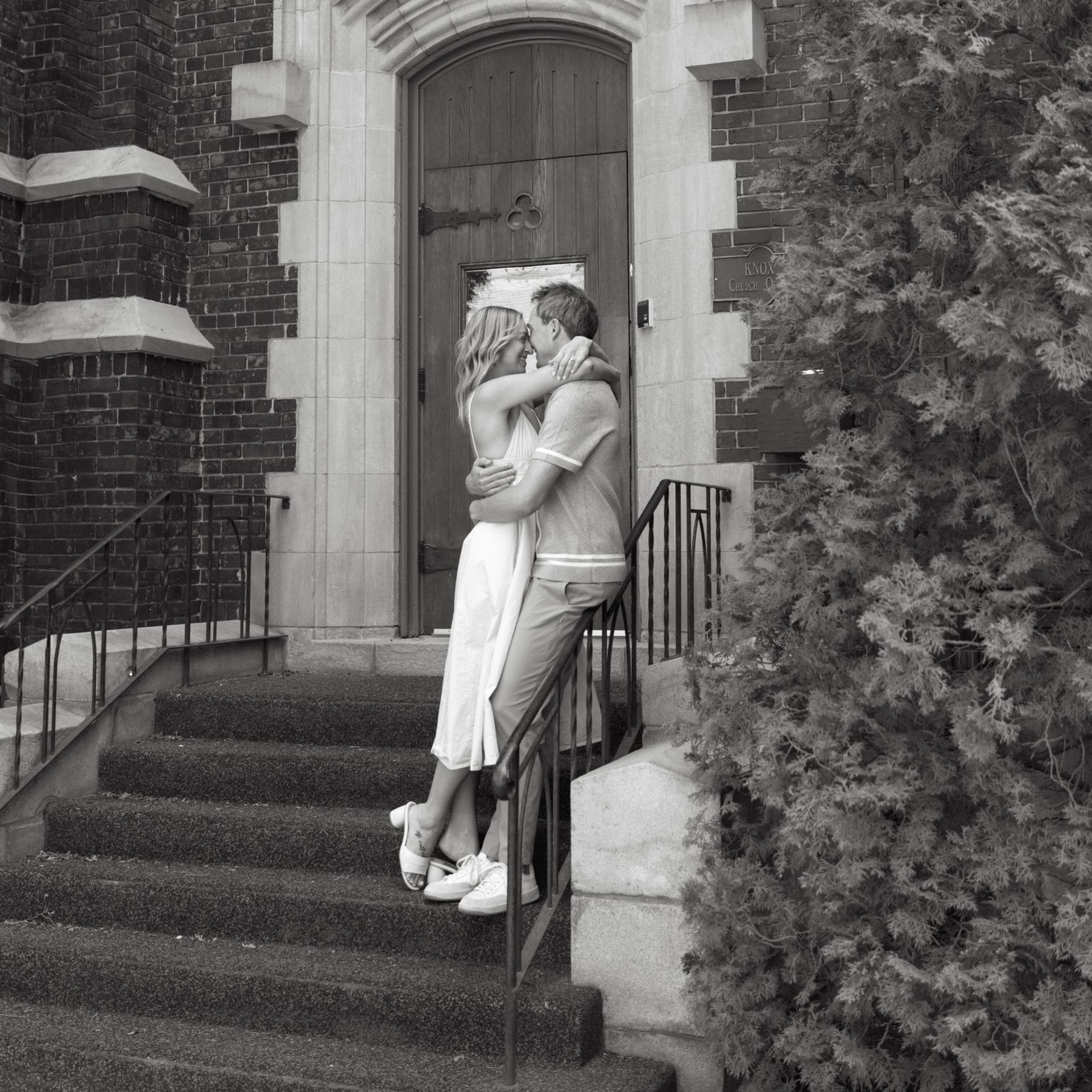 Two people embracing on stone steps outside a building entrance, surrounded by greenery.