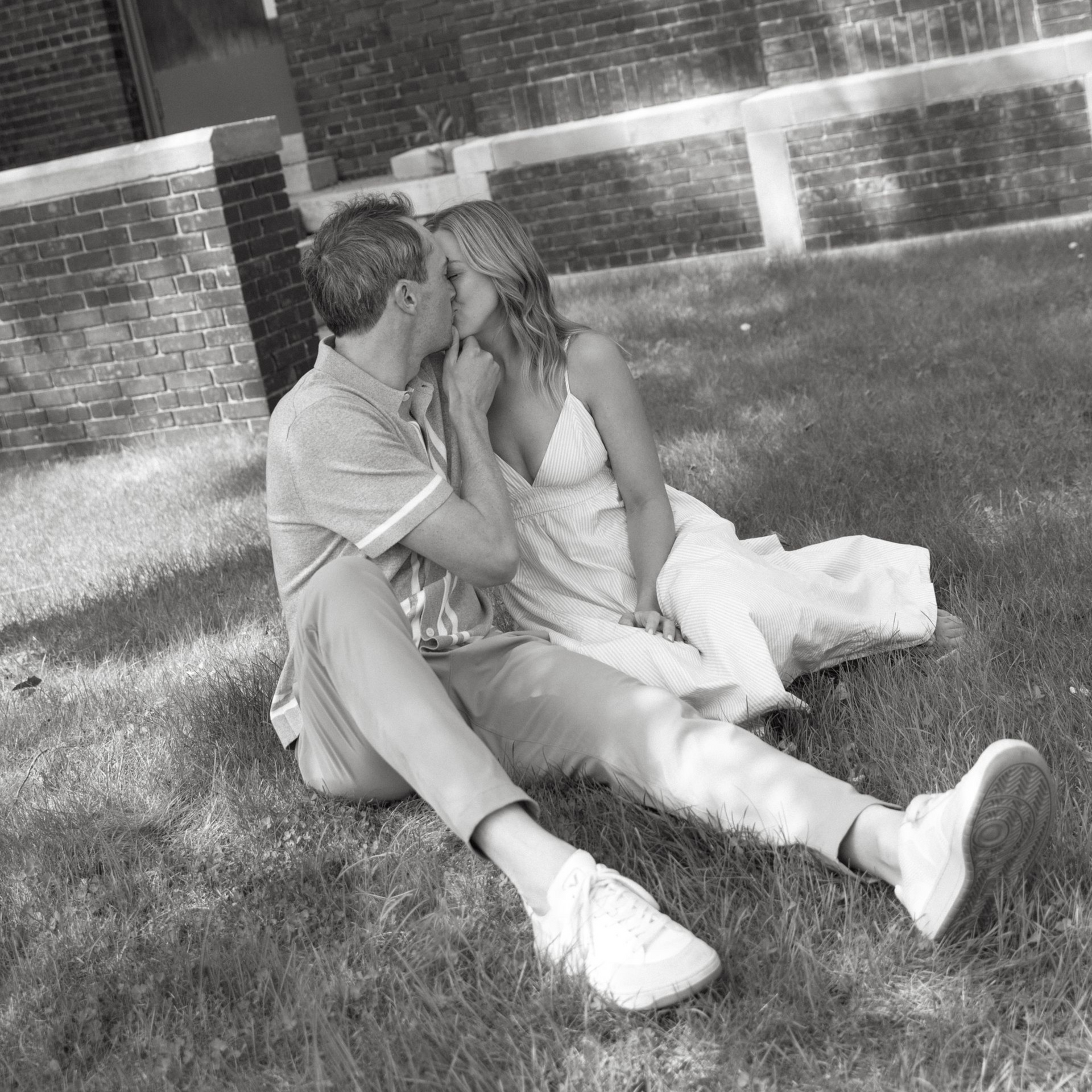 Couple sitting on grass and kissing beside a brick wall in black-and-white photo