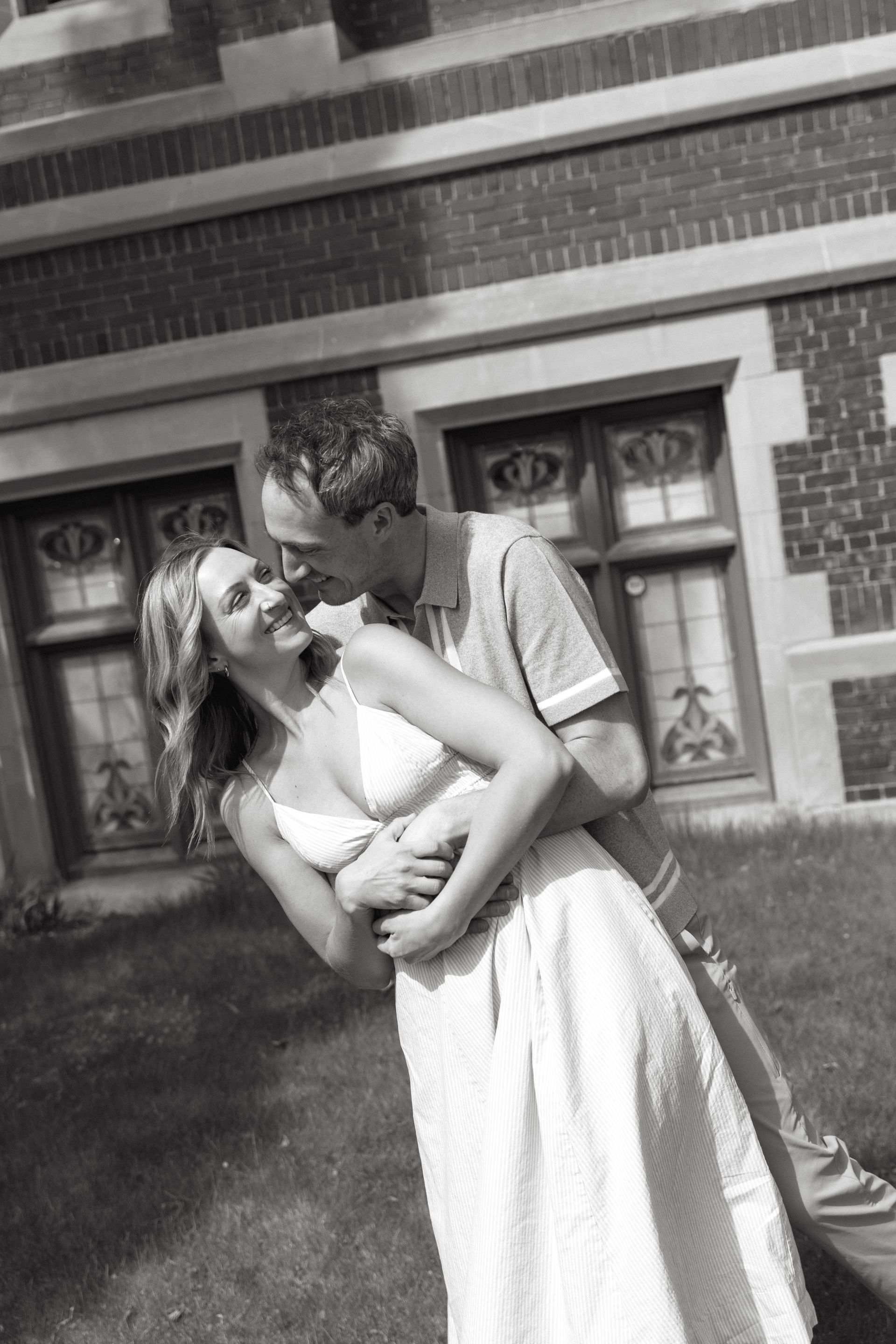Two people embracing outdoors in front of a brick building, smiling and leaning in close.