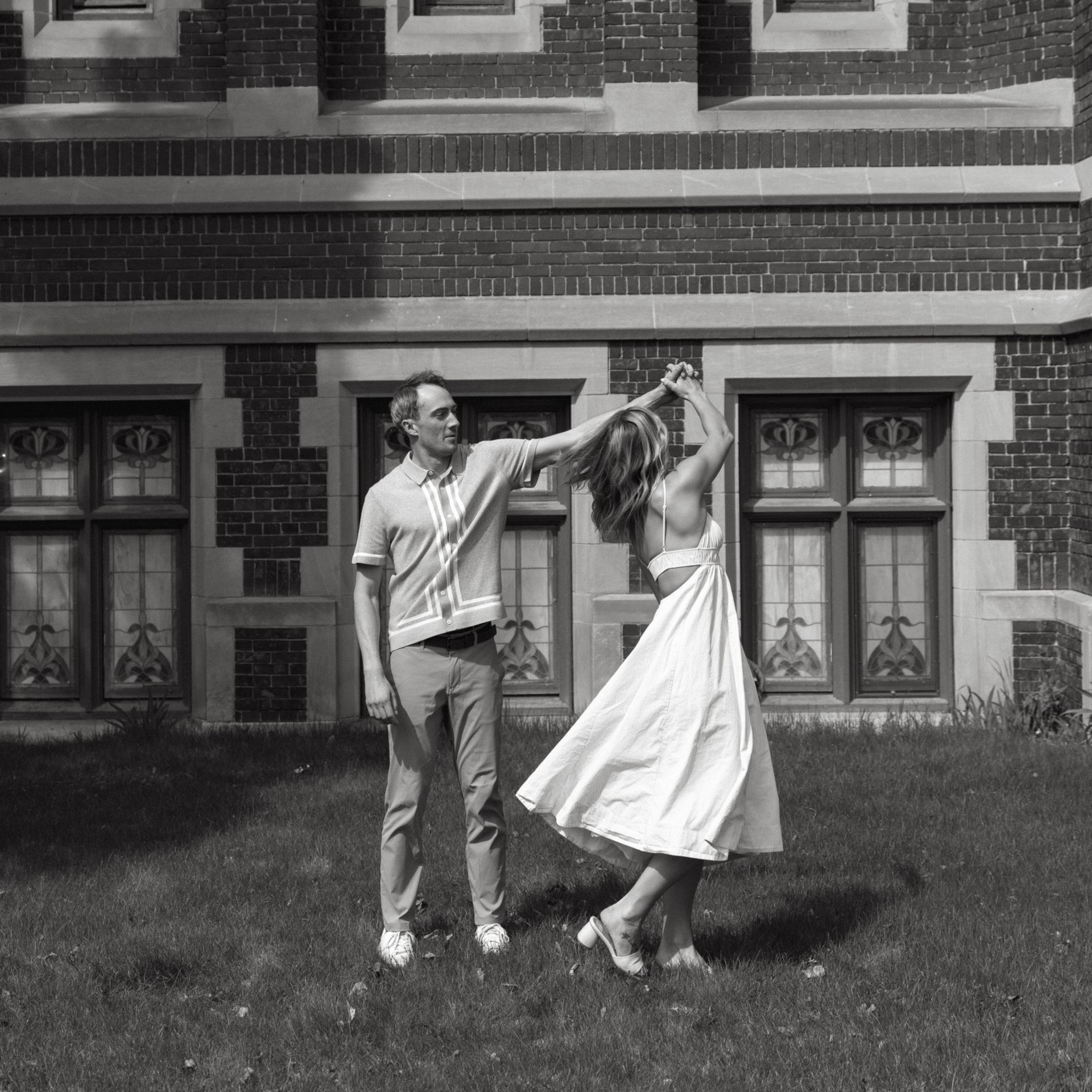 Couple dancing on a lawn in front of a brick building, with the woman twirling in a white dress.