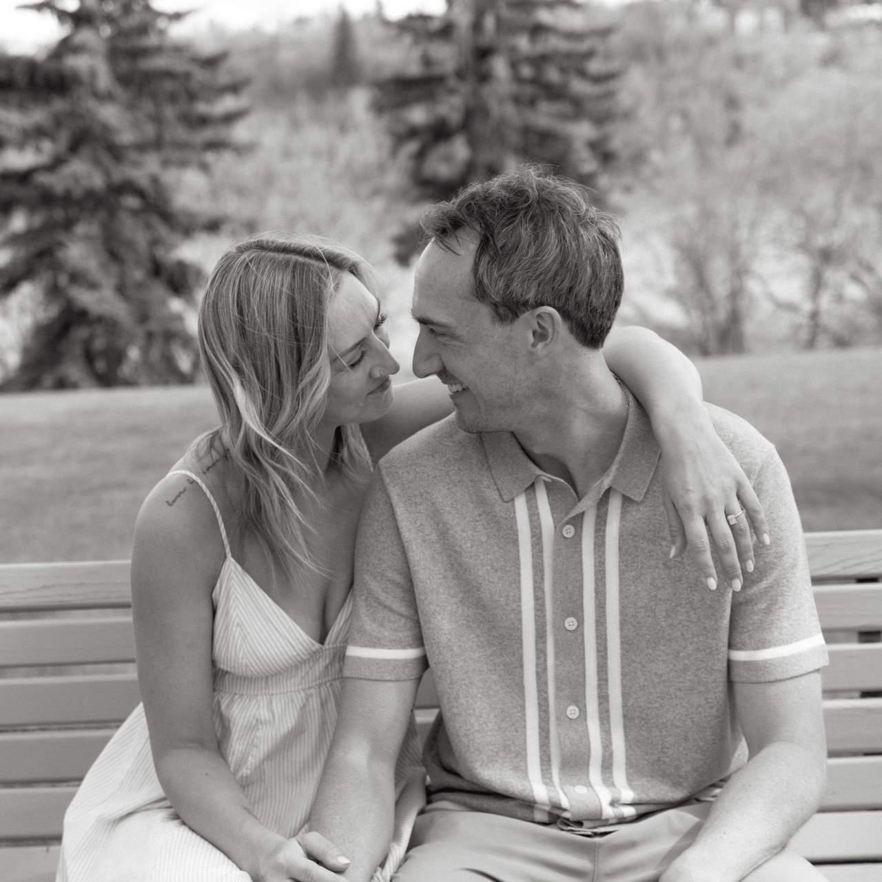 A smiling couple sits close together outdoors, leaning in and touching foreheads.