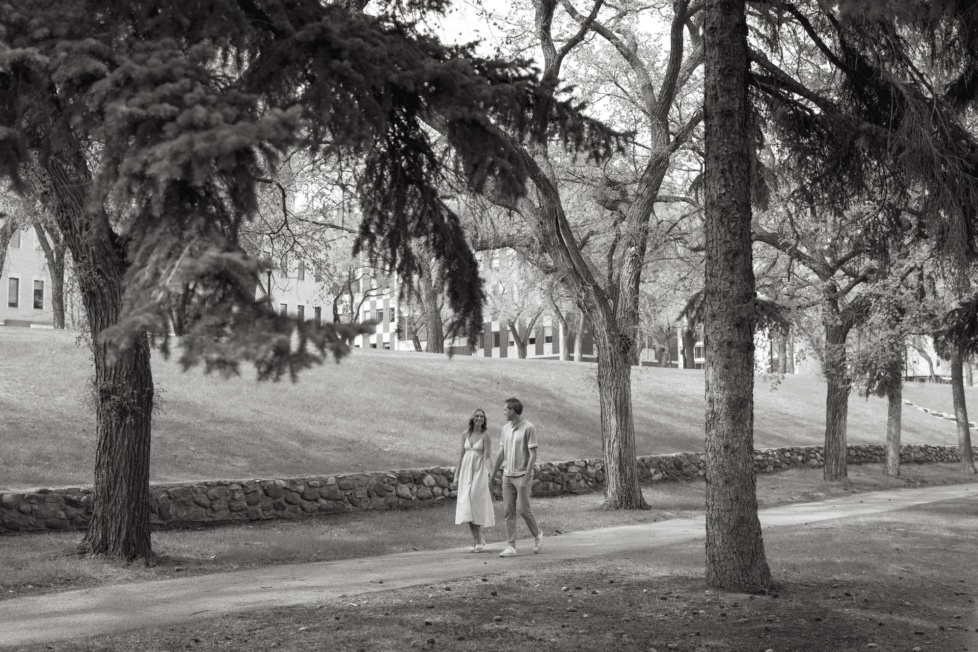 Two people walking along a tree-lined path in a park or campus, with buildings in the background.