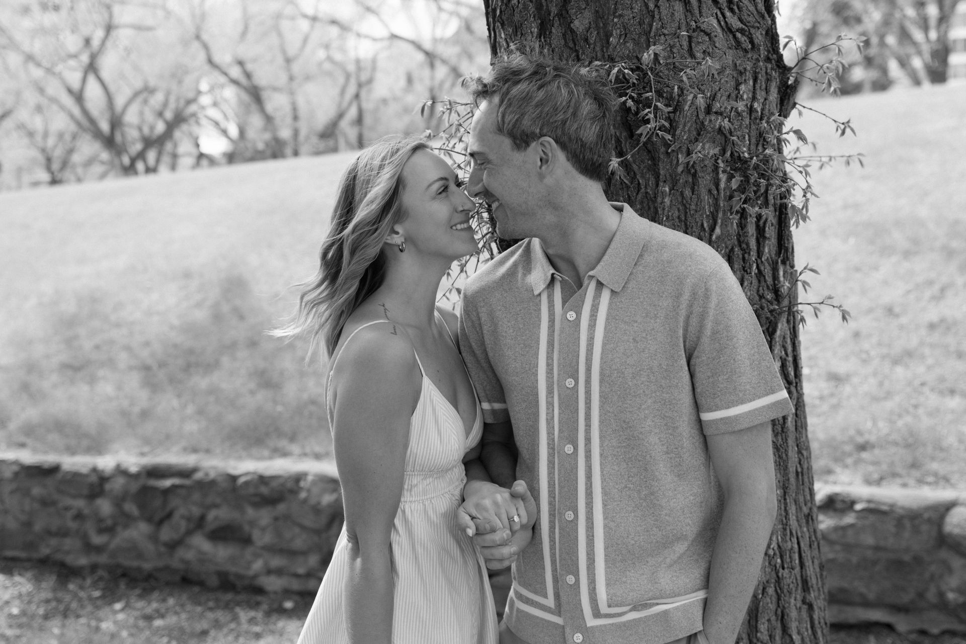 Smiling couple by a tree, leaning close in a grassy outdoor setting.