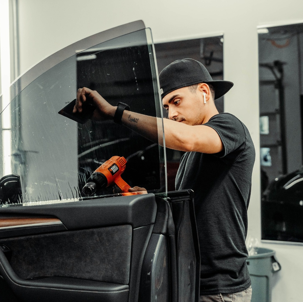 A man wearing a hat is applying window tinting to a car door.