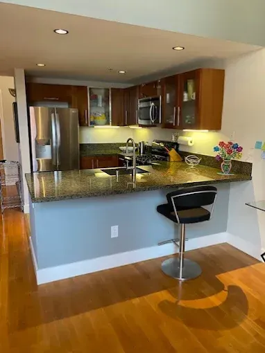 A kitchen with granite countertops, stainless steel appliances, and wood cabinets. One barstool is in front of the counter.