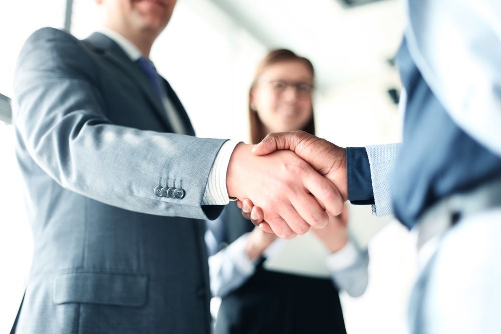 Businesspeople shaking hands in a bright office, with one man in a blue suit in the foreground.