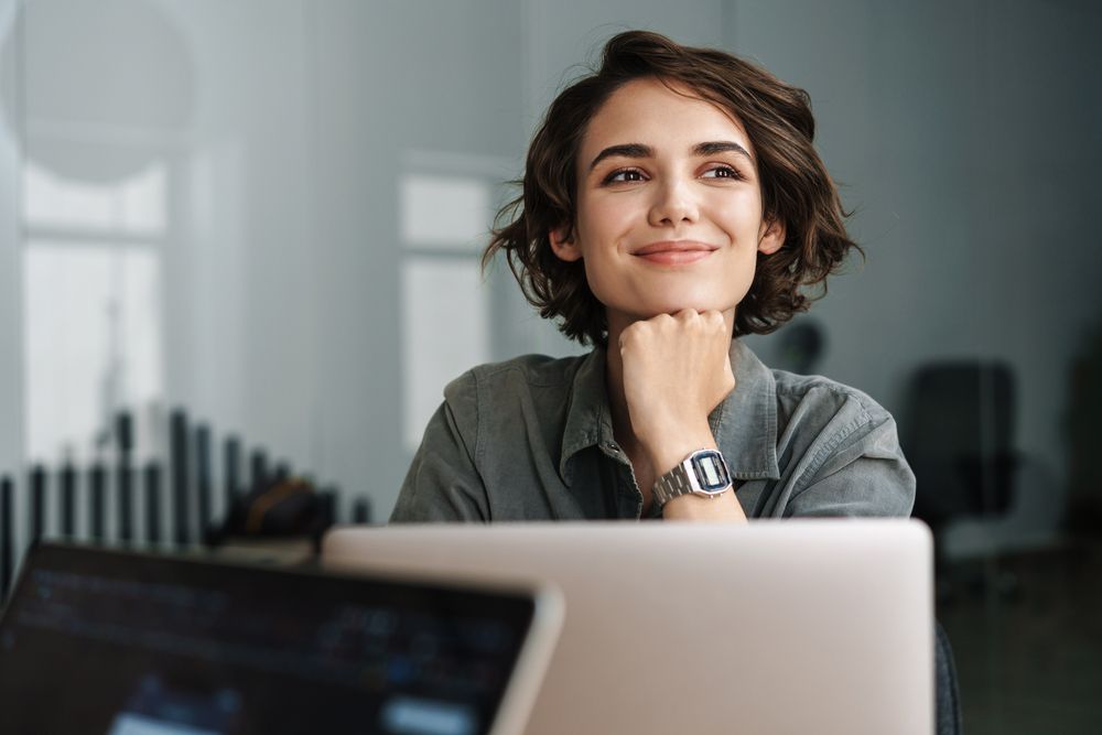 Smiling woman resting chin on hand behind a laptop in a modern office