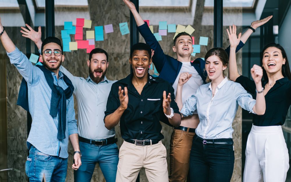 Group of coworkers celebrating with raised arms and smiles in an office with colorful sticky notes on the wall