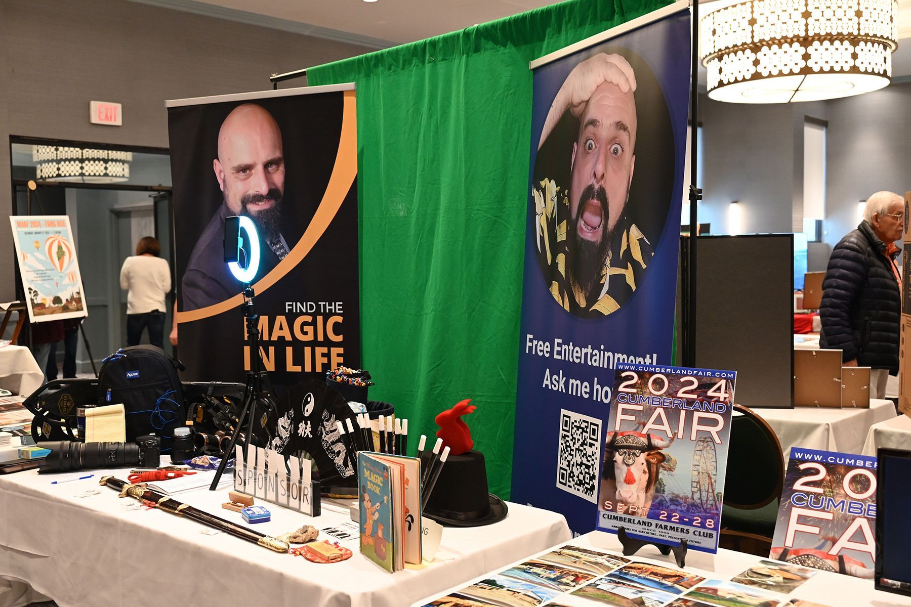 A vendor booth featuring signage for 'Magic in Life' and a 2024 fair, with a green backdrop, props, and display tables.