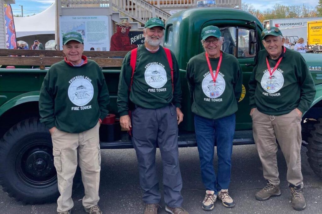 Four smiling people in matching green sweatshirts and caps stand in front of a green vintage pickup truck outdoors.