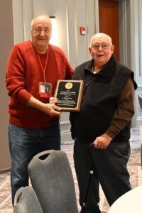 Two people stand side-by-side, one holding a plaque and the other reaching toward it, in an indoor meeting space.
