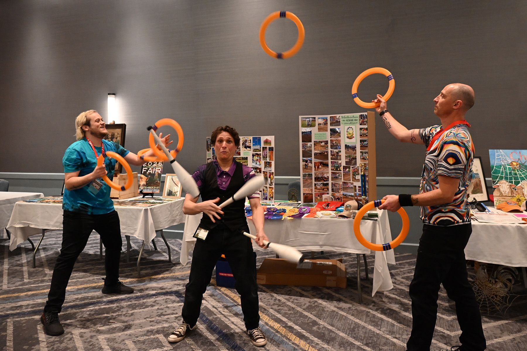 Three performers juggle hoops and clubs in a conference room with tables and display boards in the background.
