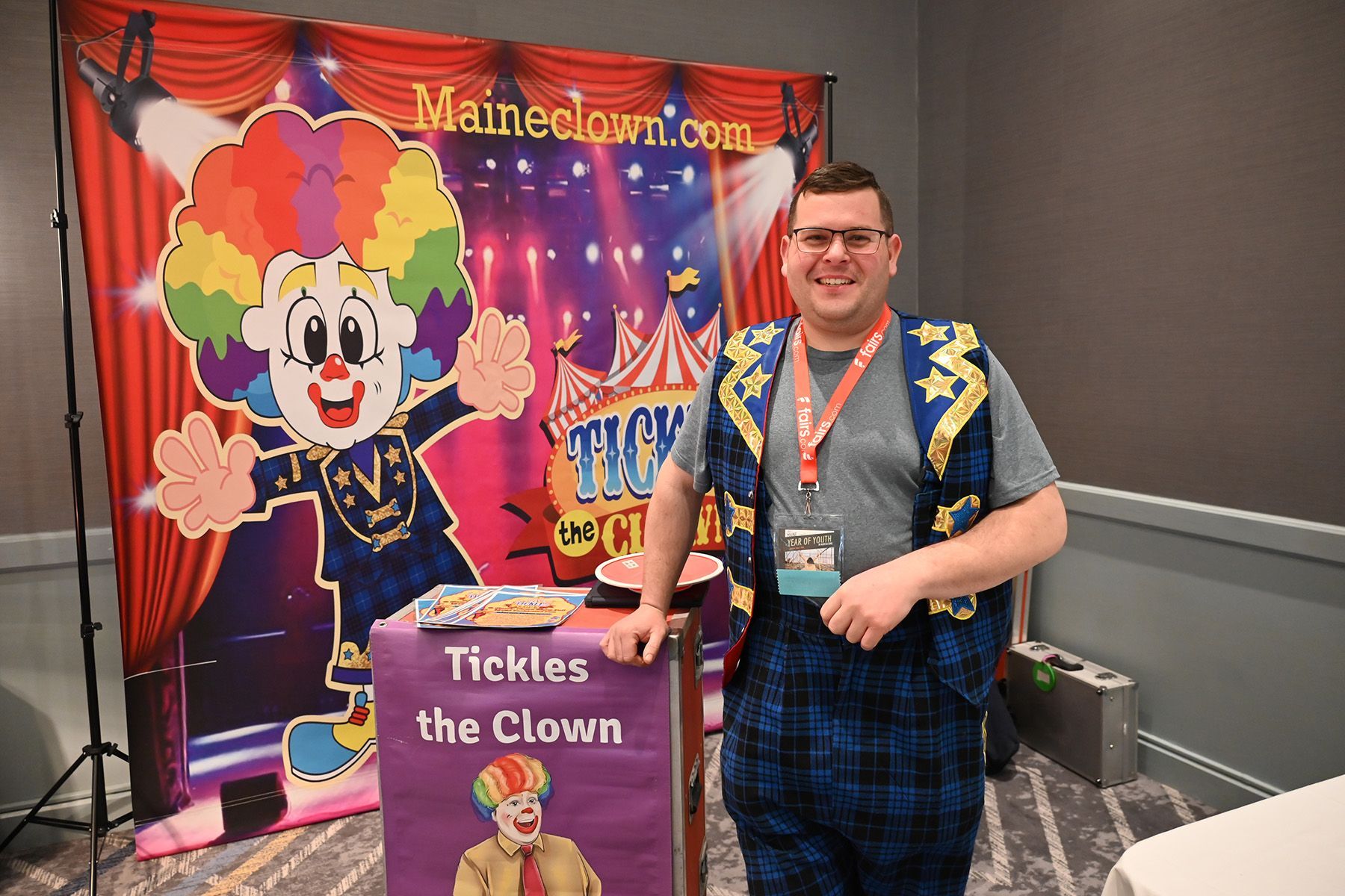 A person in a blue vest and plaid pants stands next to a 'Tickles the Clown' banner and a backdrop featuring a clown cartoon.