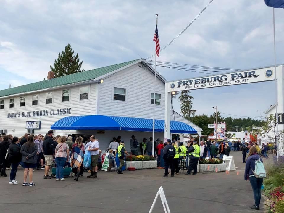 Crowd gathered at the white Fryeburg Fair entrance building under a blue awning with an American flag flying above.