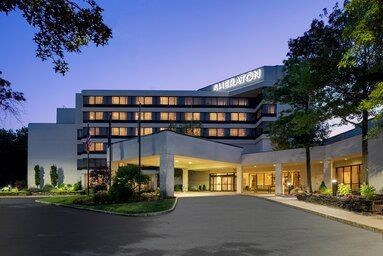 Sheraton hotel building with a large entrance canopy, paved driveway, and surrounding trees under a twilight sky.