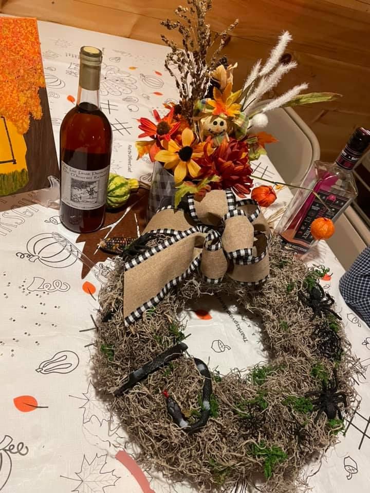 A table with an autumn-themed centerpiece, a wreath decorated with black spiders, a wine bottle, and a fall tablecloth.