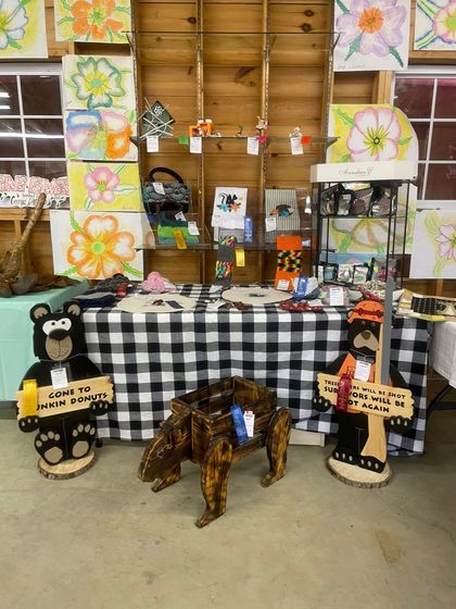 Display table at a fair with floral artwork, bear-shaped signs, and a central wooden animal sculpture on a checked cloth.