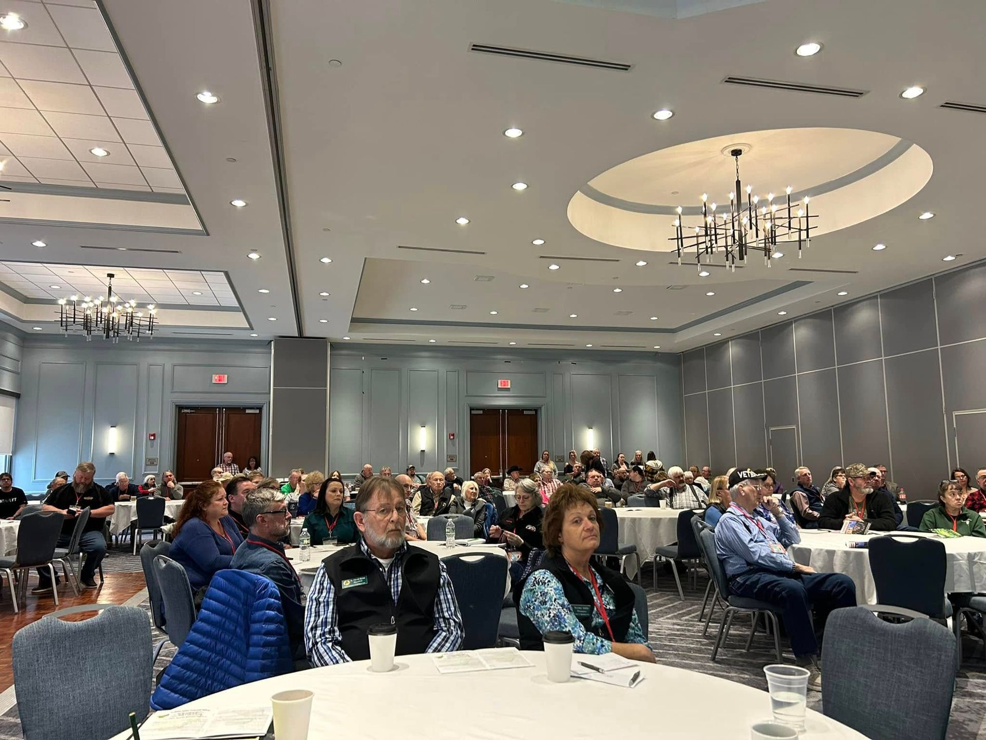 A large conference room with round tables and chairs filled with people attending a formal indoor event.
