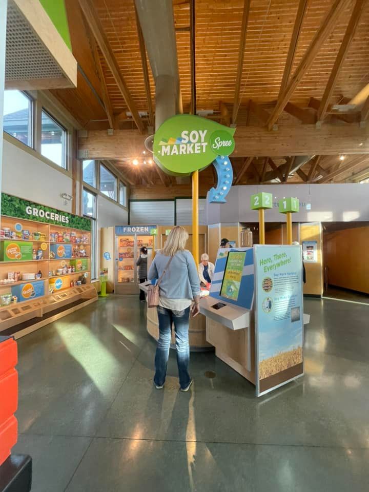 A person stands in a Soy Market exhibit featuring informative displays, product shelves, and wooden ceiling beams.