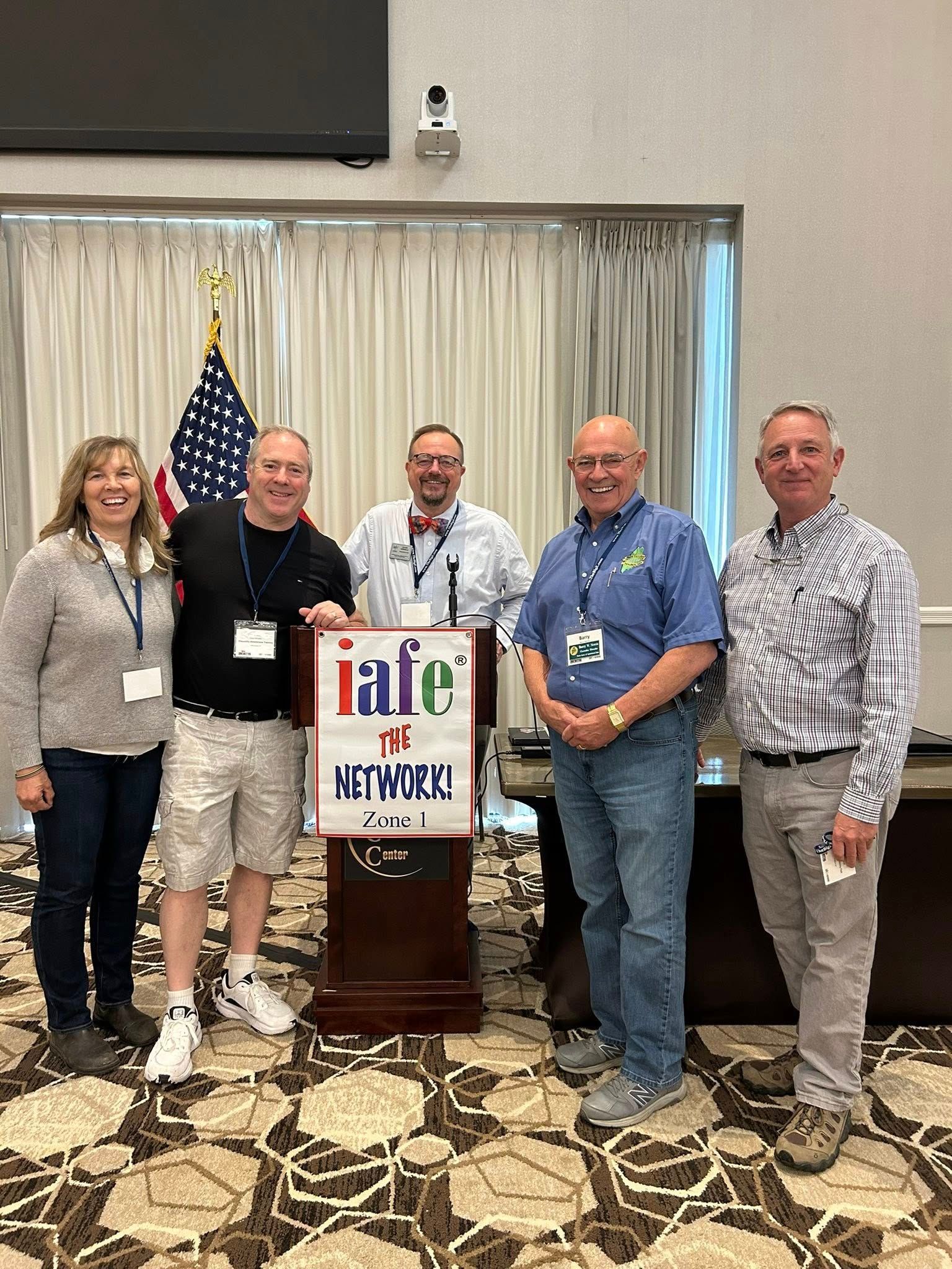 Five people stand behind a podium with an 'iafe' logo, with an American flag to the left in a conference room.