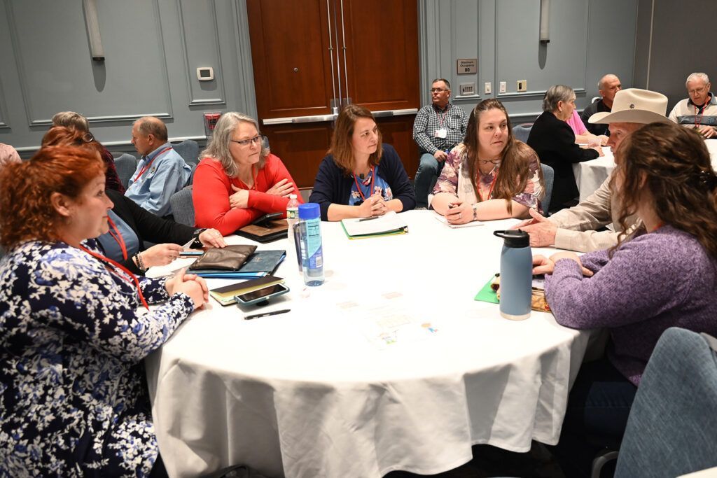 People sit around a white-covered round table at a conference, engaged in discussion with papers and water bottles.