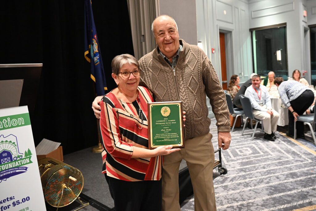 A woman and a man stand together in a ballroom, smiling while holding a framed award plaque.