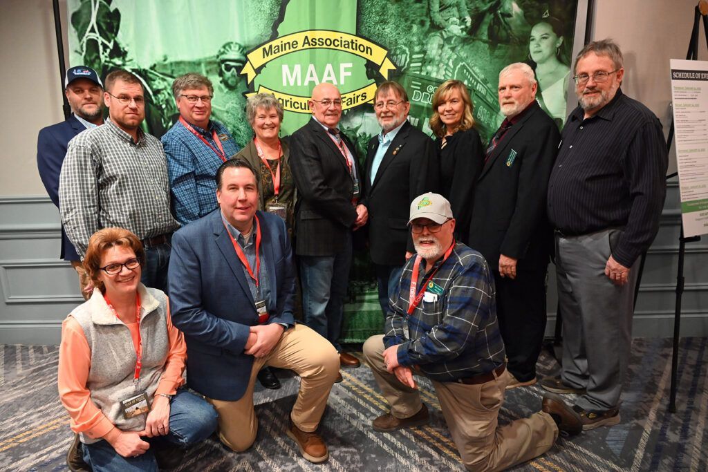 A group of people standing and kneeling in front of a green 'MAAF' event banner at an indoor gathering.