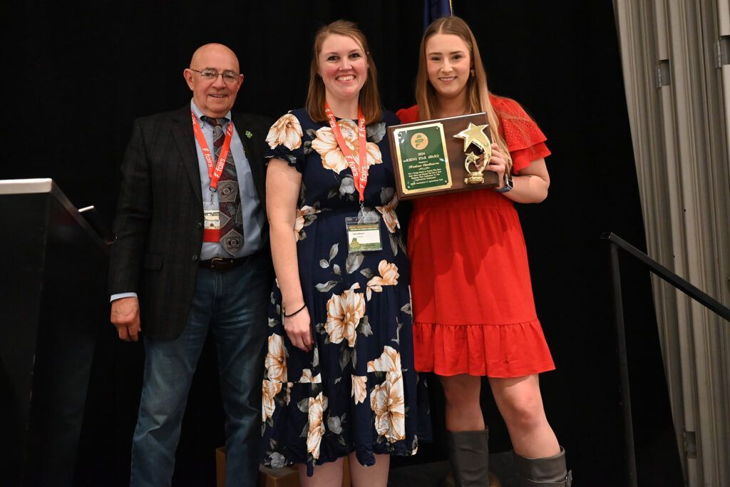 Three people stand on a stage; a person in a red dress holds an award plaque next to a person in a floral dress and a man.