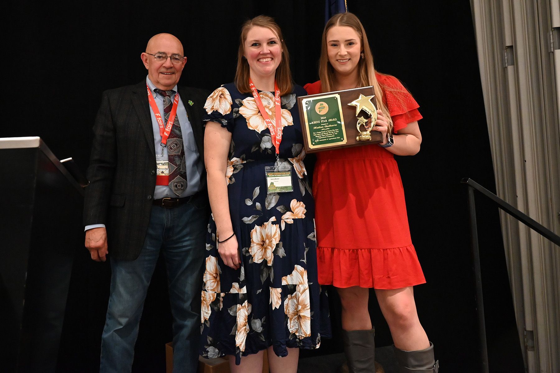 Three people stand on a stage, with the person on the right holding a plaque and trophy, all smiling at the camera.