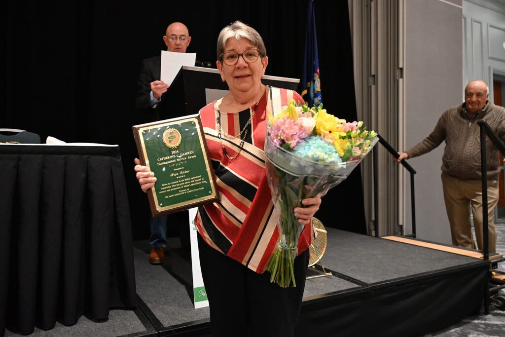 A person in a striped top holds an award plaque and a bouquet of flowers on a stage, with two people in the background.