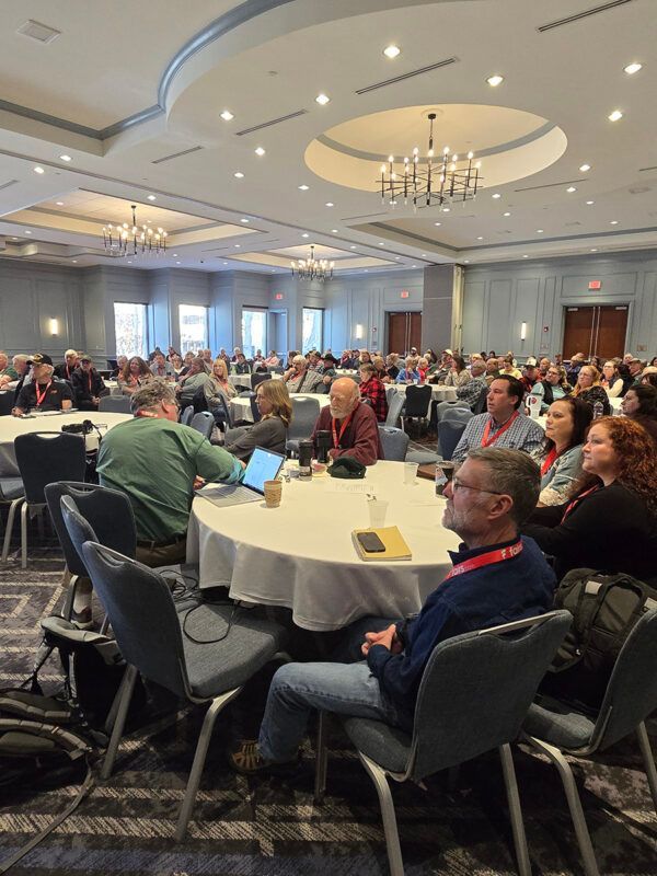 A conference room filled with people seated at round tables, listening to a presentation.