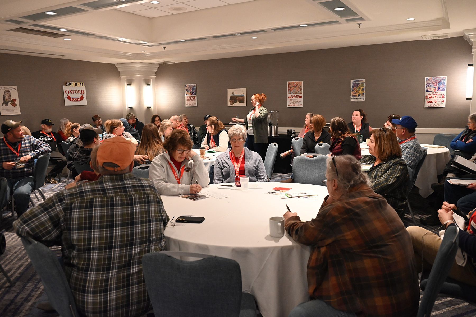 People sit at round tables in a conference room, listening to a speaker at the front. Posters are displayed on the wall.