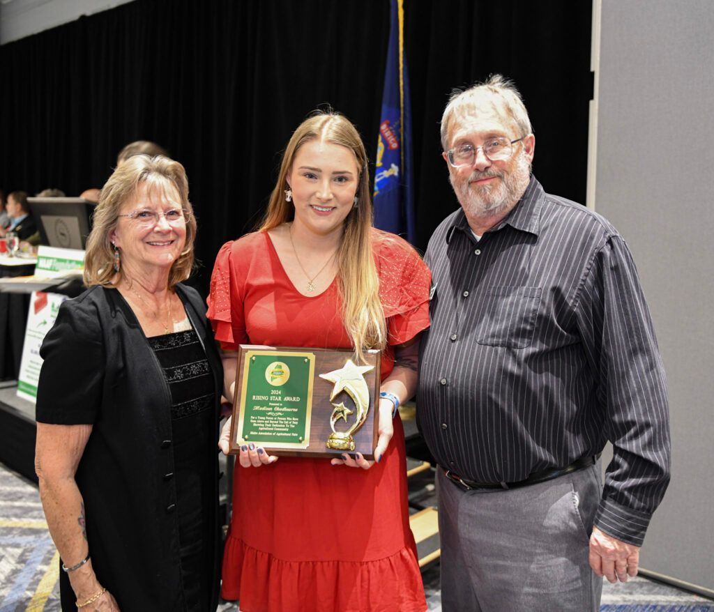 Three people stand smiling for a photo; the center person holds a commemorative plaque in an indoor, formal setting.