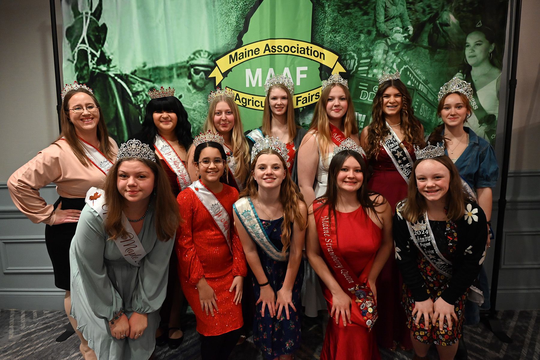 A group of people wearing sashes and tiaras pose for a photo in front of a green Maine Association of Agricultural Fairs banner.