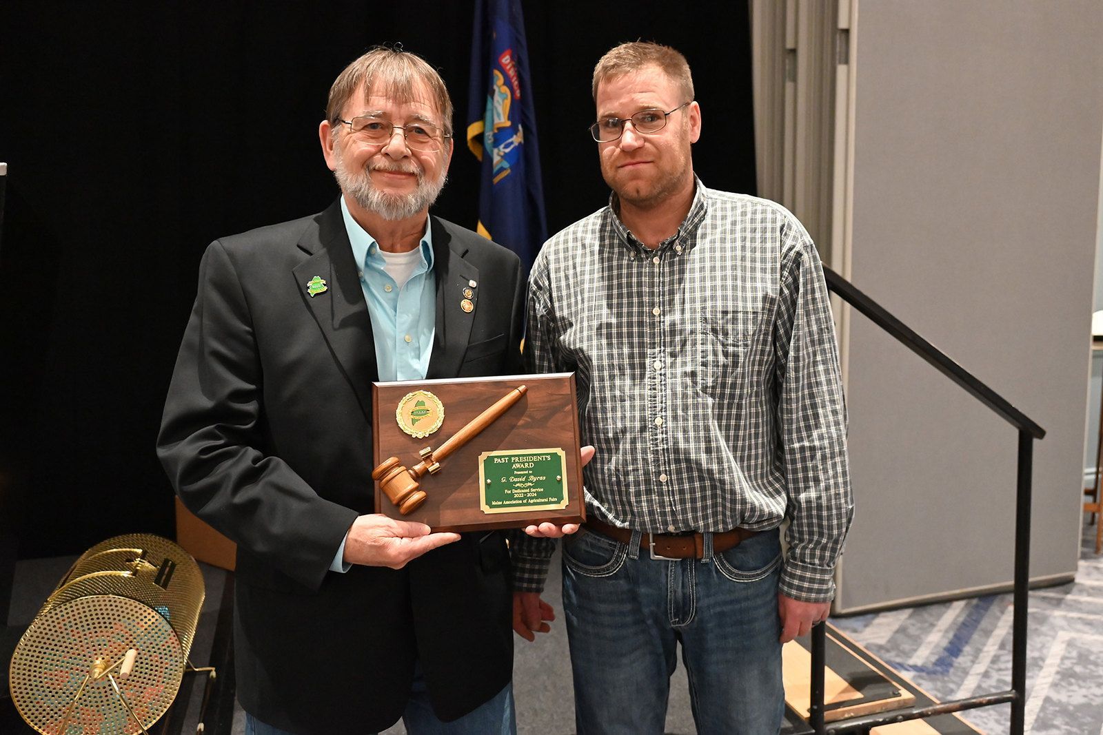 Two people stand side-by-side, one in a suit jacket, holding a wooden plaque featuring a gavel and an engraved plate.