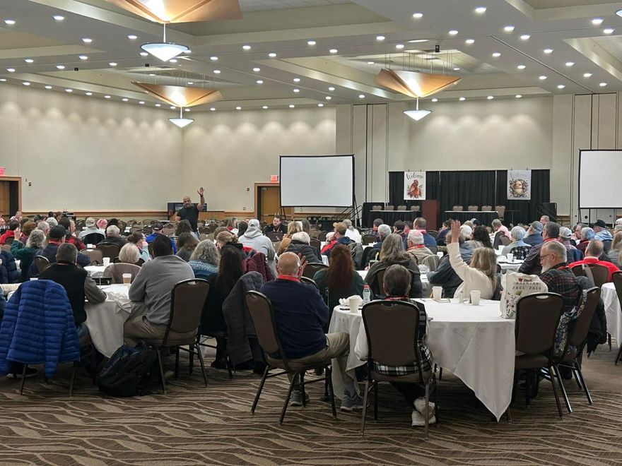 A large, indoor conference room filled with people seated at round tables, some facing forward toward two projection screens.