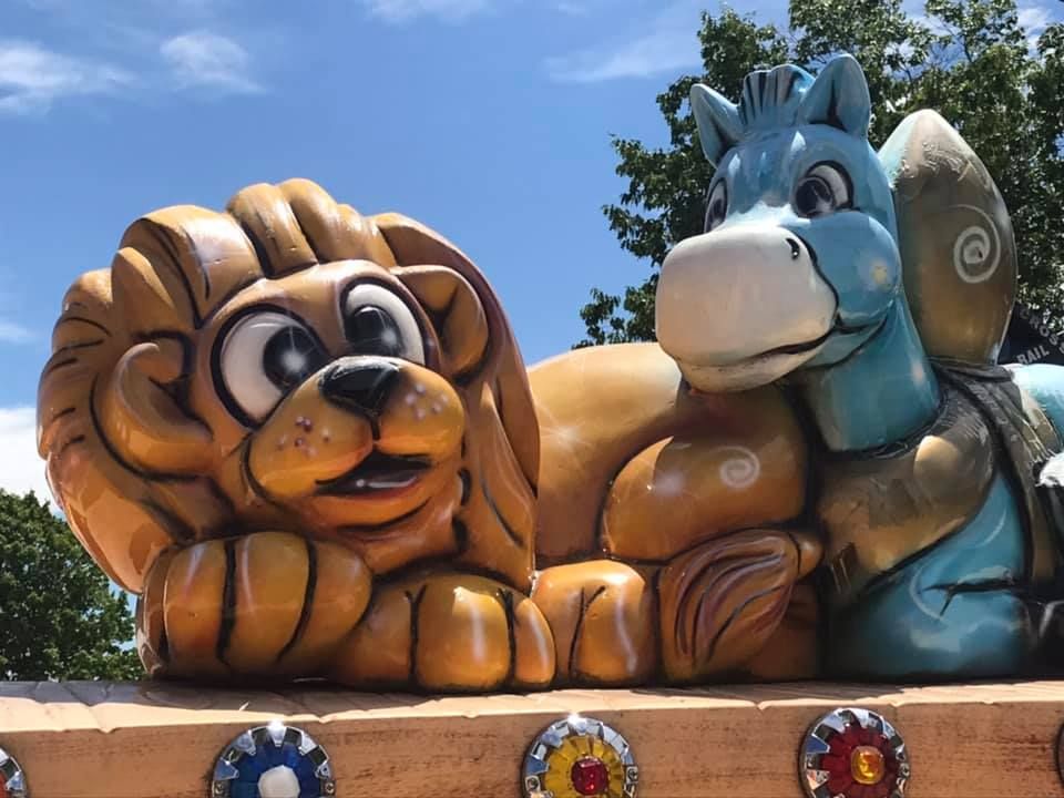 Cartoonish, carved wood sculptures of a brown lion and a light blue horse side-by-side against a blue sky.
