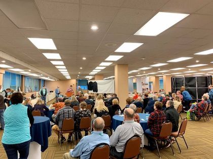 People sit at round tables in a large room for an indoor event, with a speaker addressing the audience at the front.