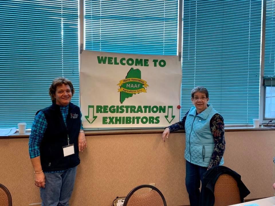 Two people stand in front of a white sign reading 'Welcome to Registration Exhibitors' inside a room with window blinds.