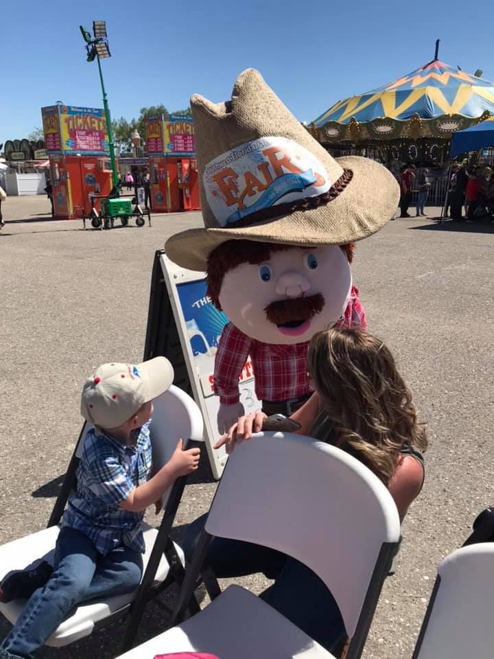 A mascot wearing a large cowboy hat greets a child sitting in a chair at a fairground with a carousel in the background.