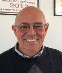 A smiling person wearing glasses and a dark sweater, positioned in front of two framed documents.