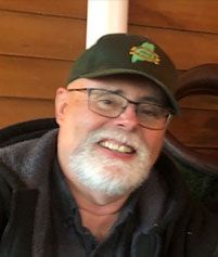 A smiling person with a white beard and glasses, wearing a dark cap and jacket, in front of a wood-paneled wall.