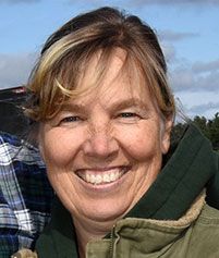 A person with shoulder-length blonde hair and a warm smile, wearing a green hooded jacket outdoors.