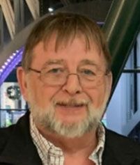 A person with a beard and glasses smiles while wearing a collared, patterned shirt under a dark jacket.