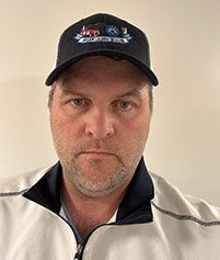 A person with stubble wears a dark-colored baseball cap and a white quarter-zip jacket against a plain wall.