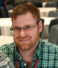 A person with short brown hair, glasses, and a beard, wearing a green plaid button-down shirt in a room with white tables.
