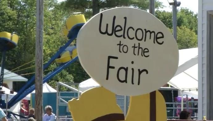 A sign that says 'Welcome to the Fair' in front of a carnival ride with yellow cars.