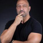 A man with a shaved head and facial hair, wearing a black t-shirt, poses with one hand near his chin against a black backdrop.