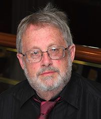 A person with a beard and glasses wearing a black shirt and a maroon tie looks toward the camera.